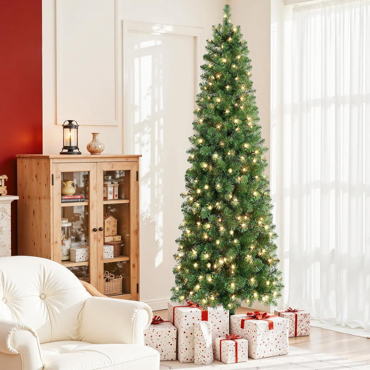 Lit green pencil Christmas tree beside a bright window and white armchair.