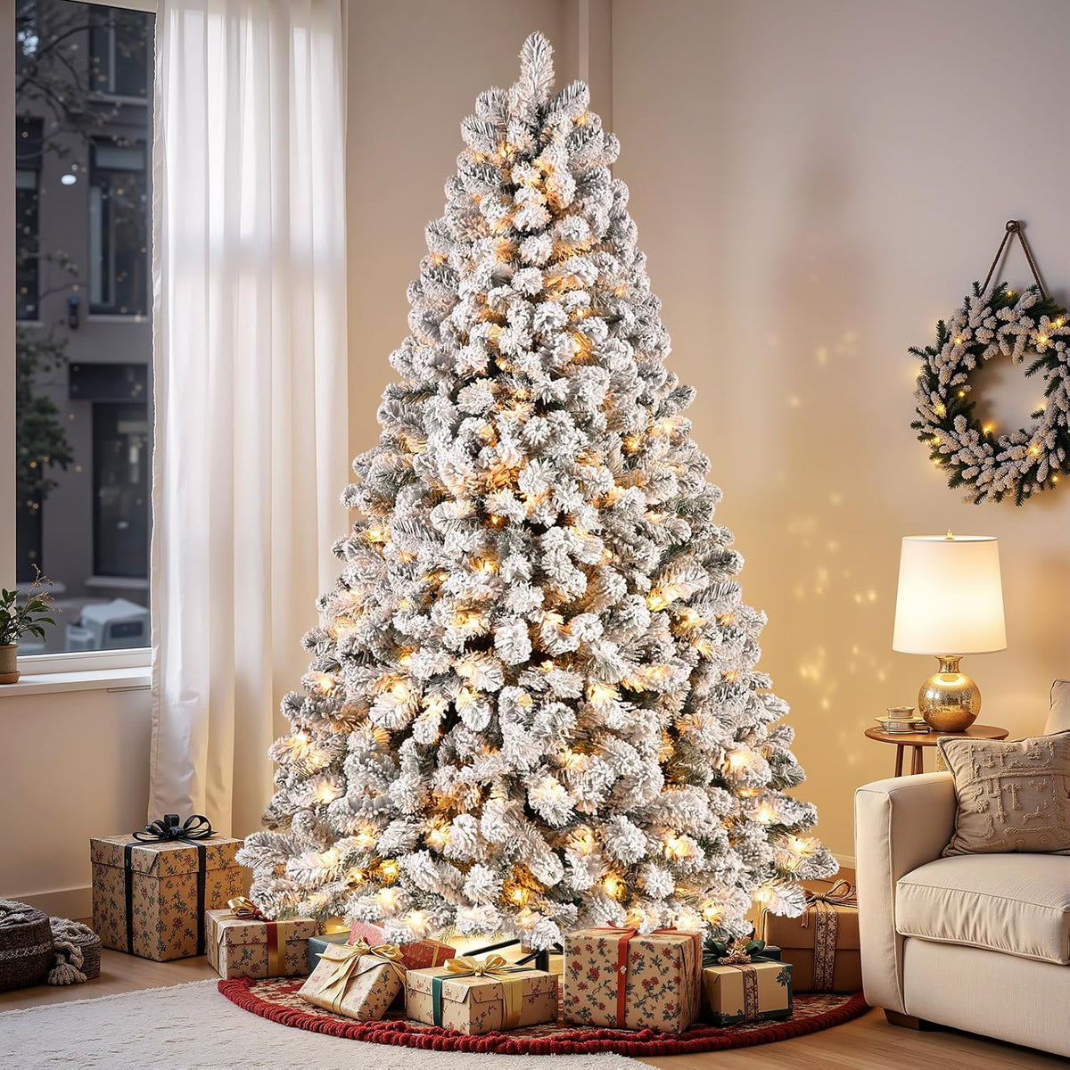 Lit snow-flocked spruce Christmas tree beside a living-room window, sofa and gifts.